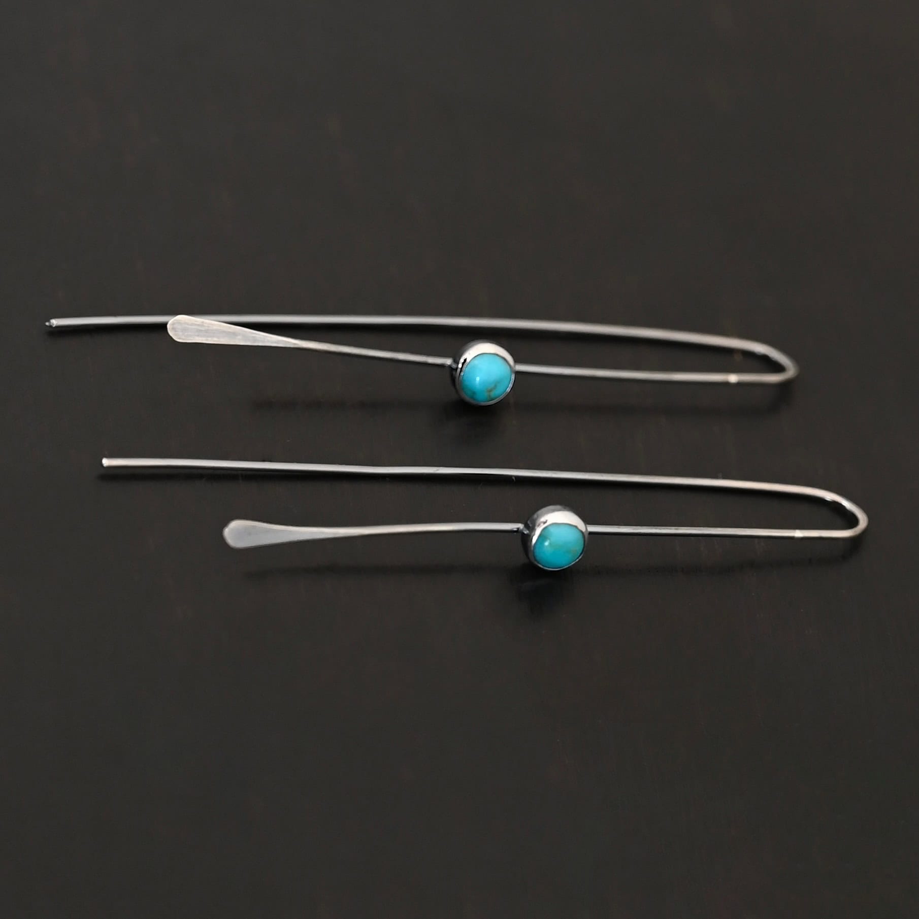 Nimmer Earrings "Nimmer" Earrings. Each earring is a long, thin line of curved silver at the top, with a hammered paddle end. Each earring has a small, circular turquoise in the center of the line on the front.