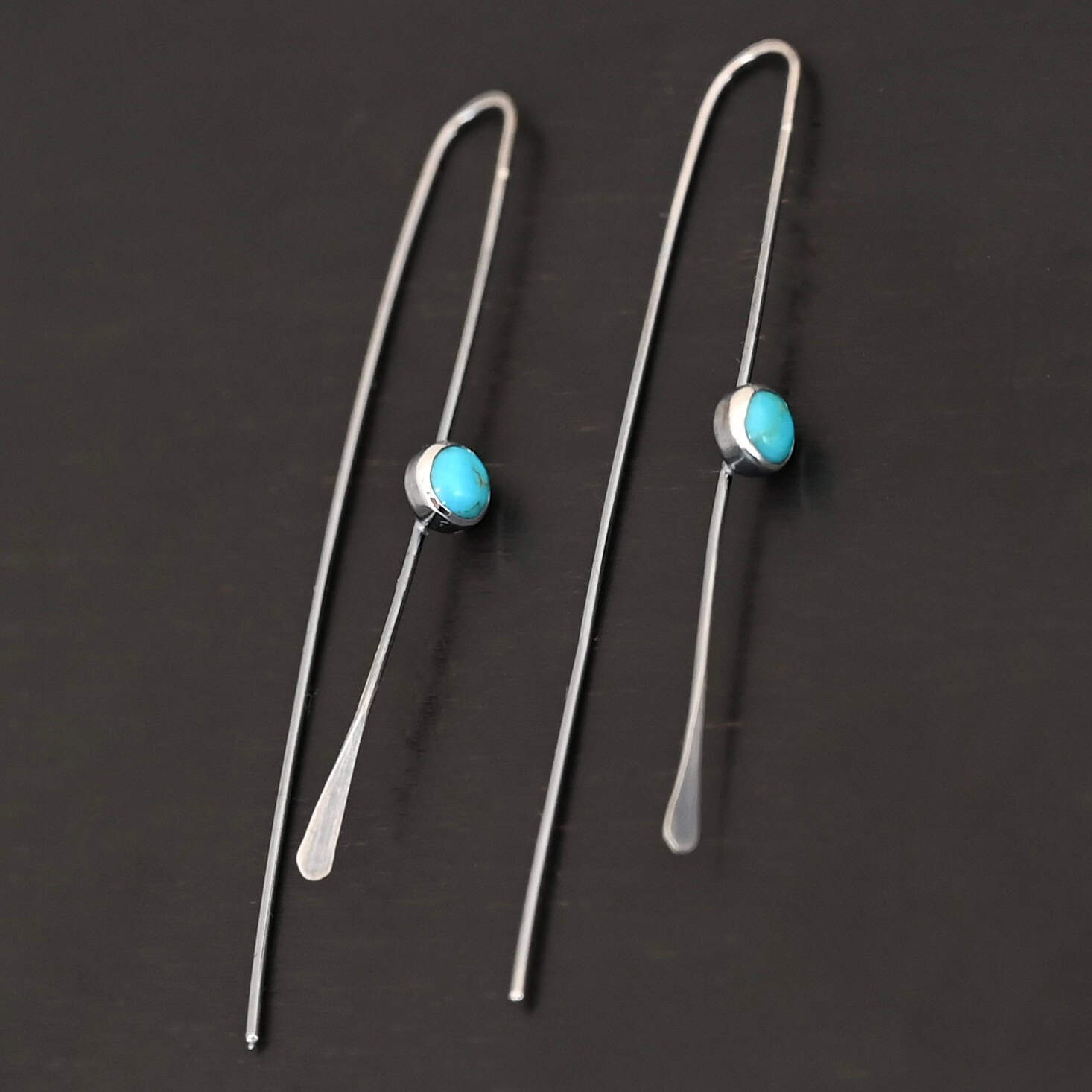 Nimmer Earrings "Nimmer" Earrings. Each earring is a long, thin line of curved silver at the top, with a hammered paddle end. Each earring has a small, circular turquoise in the center of the line on the front.