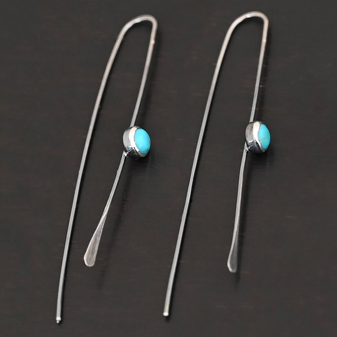 Nimmer Earrings "Nimmer" Earrings. Each earring is a long, thin line of curved silver at the top, with a hammered paddle end. Each earring has a small, circular turquoise in the center of the line on the front.