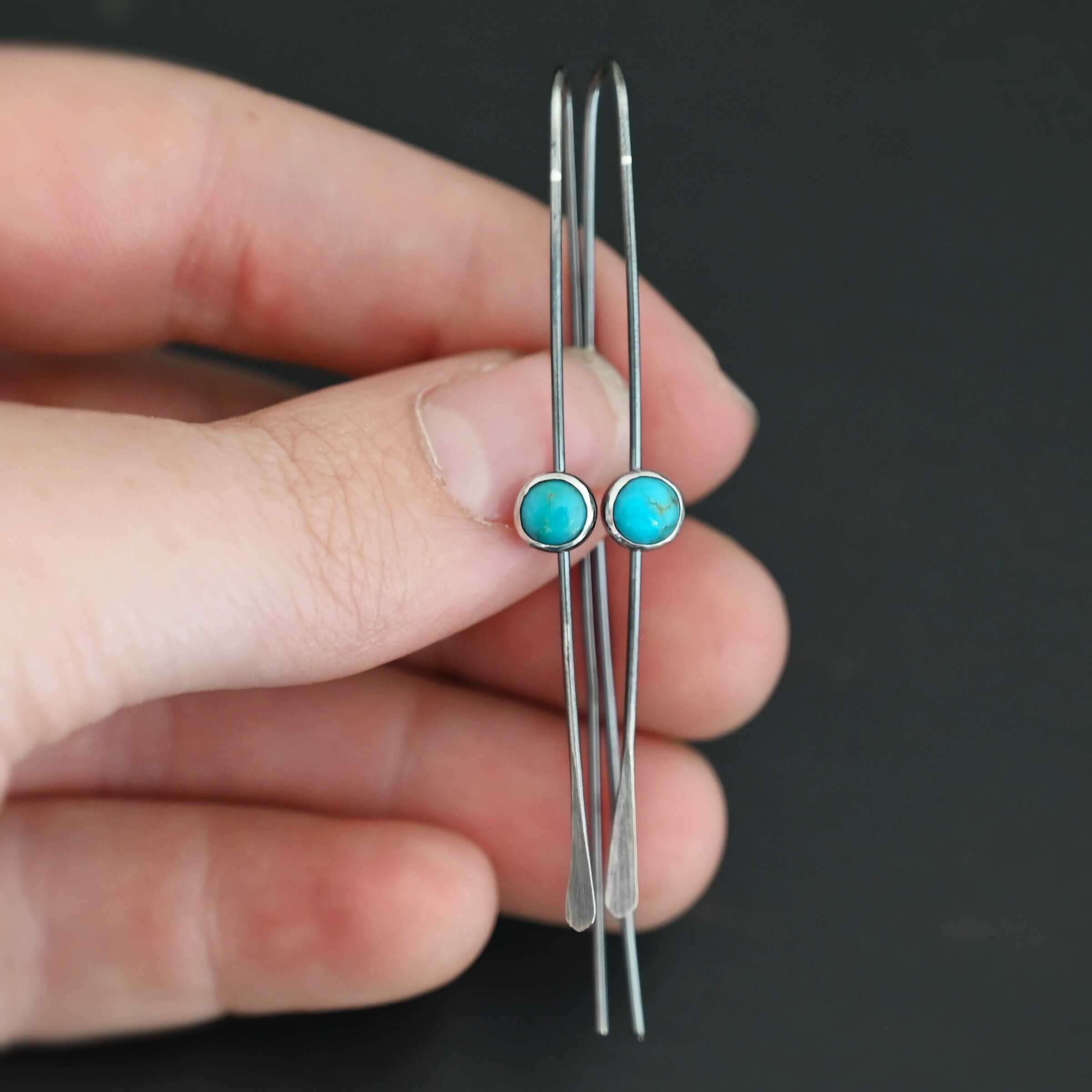 Nimmer Earrings "Nimmer" Earrings. Each earring is a long, thin line of curved silver at the top, with a hammered paddle end. Each earring has a small, circular turquoise in the center of the line on the front.
