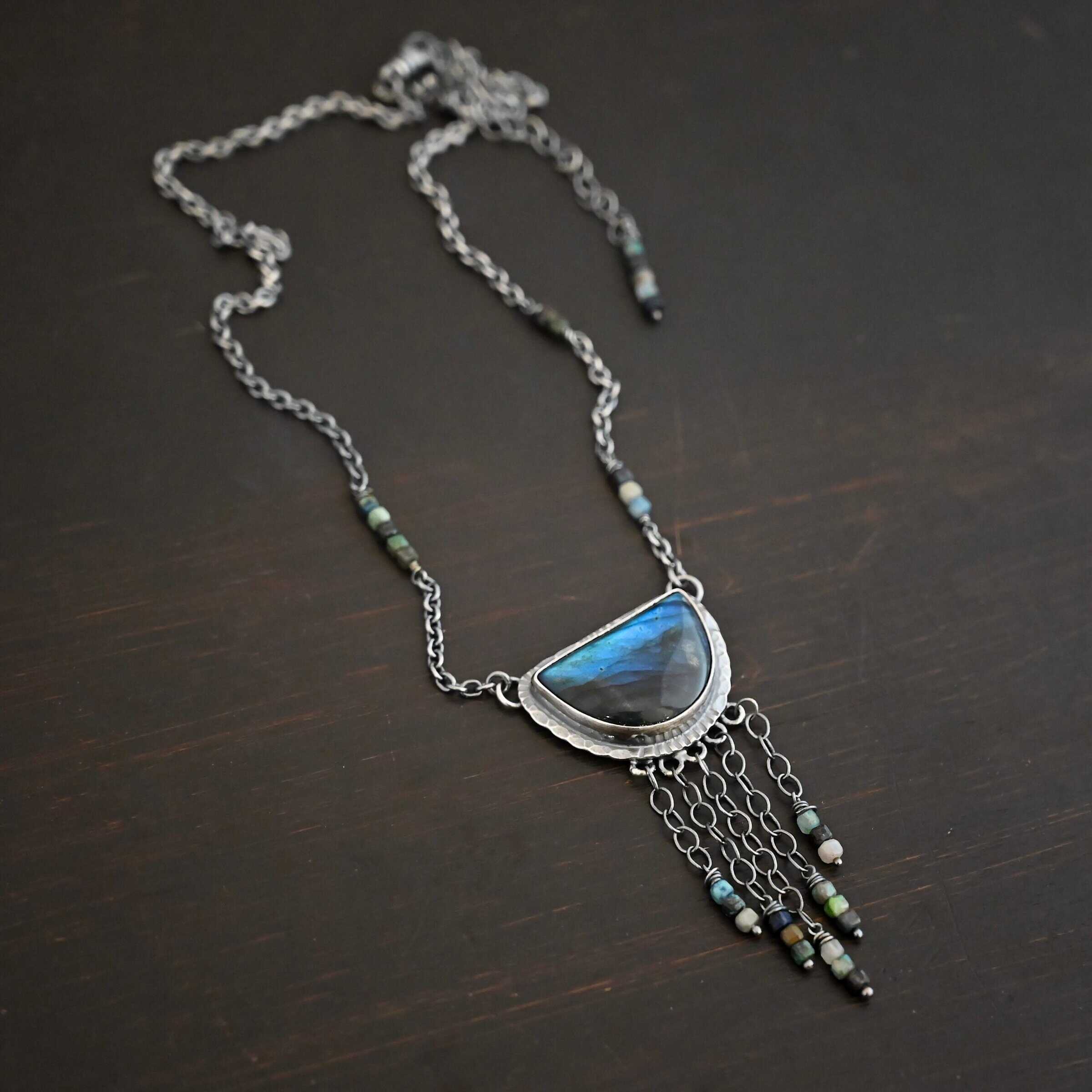 Dream Catcher Necklace Close up of the "Dream Catcher" Necklace. Made out of sterling silver, labradorite, and chrysocolla beads. The labradorite is a half circle shape, with the flat half at the top from where the circle would be cut in half. There are five short strands of silver chain with chrysocolla beads on each end of the chains. The labradorite is set in hammered sterling silver and on each end of the top of the setting is a loop. Attached to the loops are silver chain with chrysocolla beads on them. Necklace is laying against a dark background.