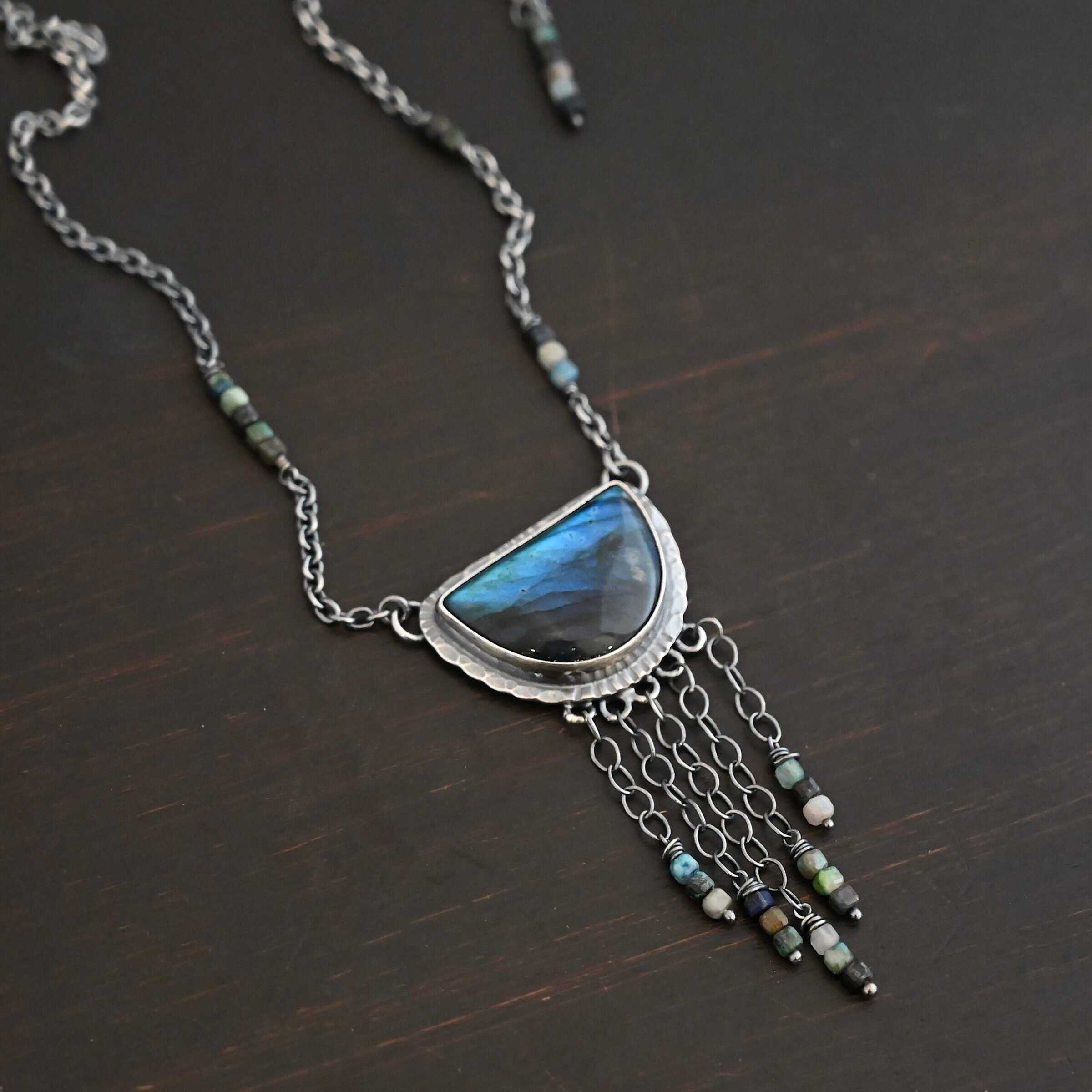 Dream Catcher Necklace Close up of the "Dream Catcher" Necklace. Made out of sterling silver, labradorite, and chrysocolla beads. The labradorite is a half circle shape, with the flat half at the top from where the circle would be cut in half. There are five short strands of silver chain with chrysocolla beads on each end of the chains. The labradorite is set in hammered sterling silver and on each end of the top of the setting is a loop. Attached to the loops are silver chain with chrysocolla beads on them. Necklace is laying against a dark background.