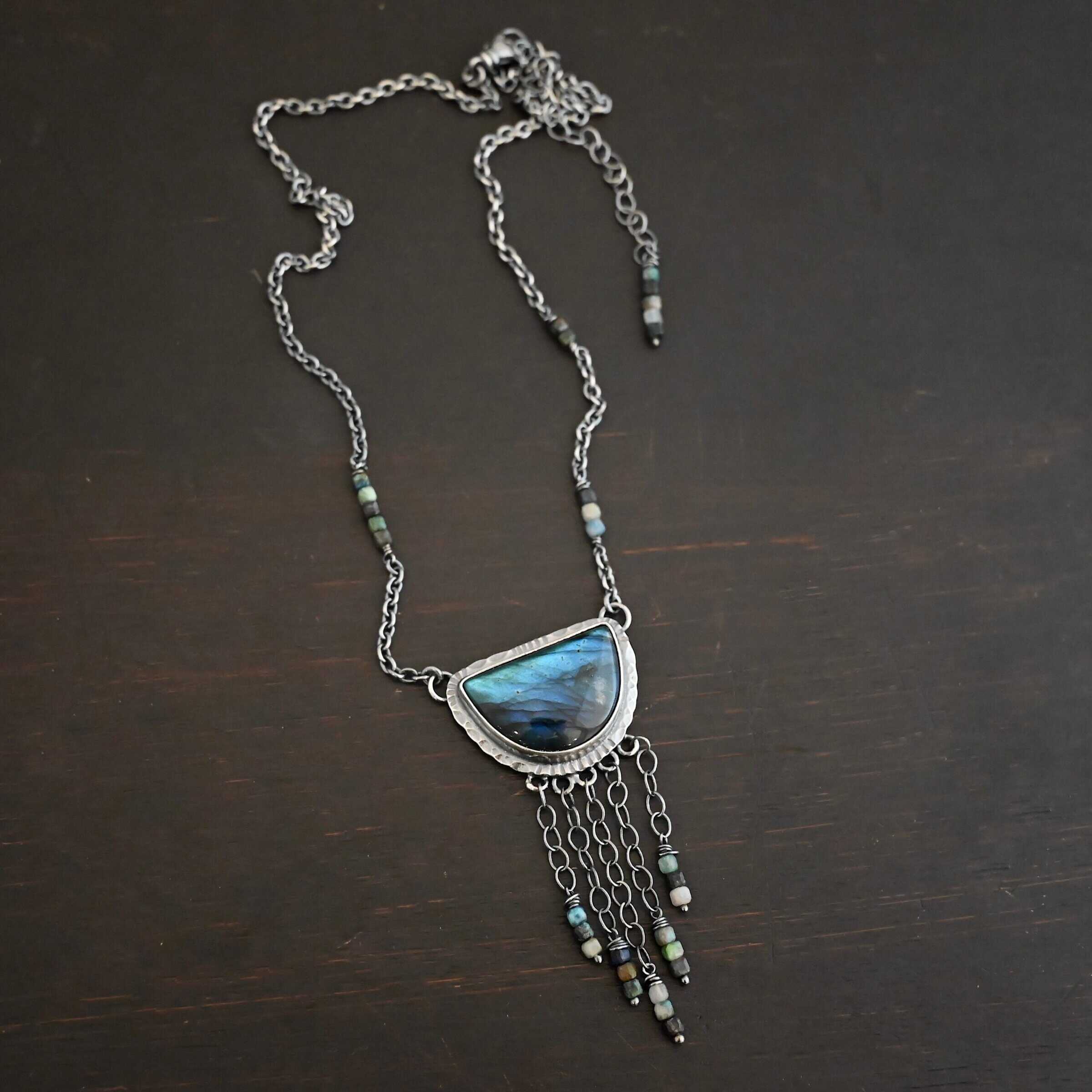 Dream Catcher Necklace Close up of the "Dream Catcher" Necklace. Made out of sterling silver, labradorite, and chrysocolla beads. The labradorite is a half circle shape, with the flat half at the top from where the circle would be cut in half. There are five short strands of silver chain with chrysocolla beads on each end of the chains. The labradorite is set in hammered sterling silver and on each end of the top of the setting is a loop. Attached to the loops are silver chain with chrysocolla beads on them. Necklace is laying against a dark background.