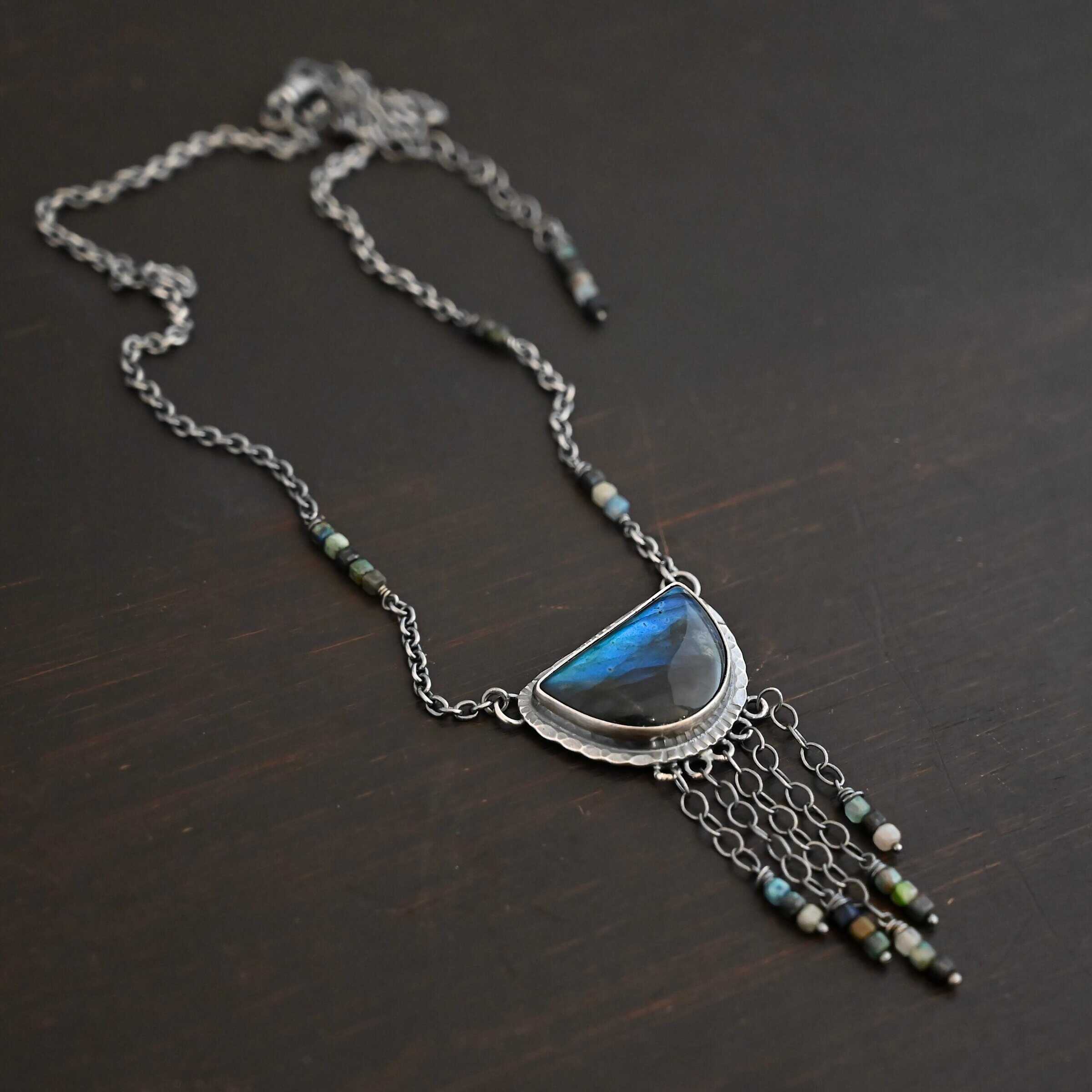 Dream Catcher Necklace Close up of the "Dream Catcher" Necklace. Made out of sterling silver, labradorite, and chrysocolla beads. The labradorite is a half circle shape, with the flat half at the top from where the circle would be cut in half. There are five short strands of silver chain with chrysocolla beads on each end of the chains. The labradorite is set in hammered sterling silver and on each end of the top of the setting is a loop. Attached to the loops are silver chain with chrysocolla beads on them. Necklace is laying against a dark background.