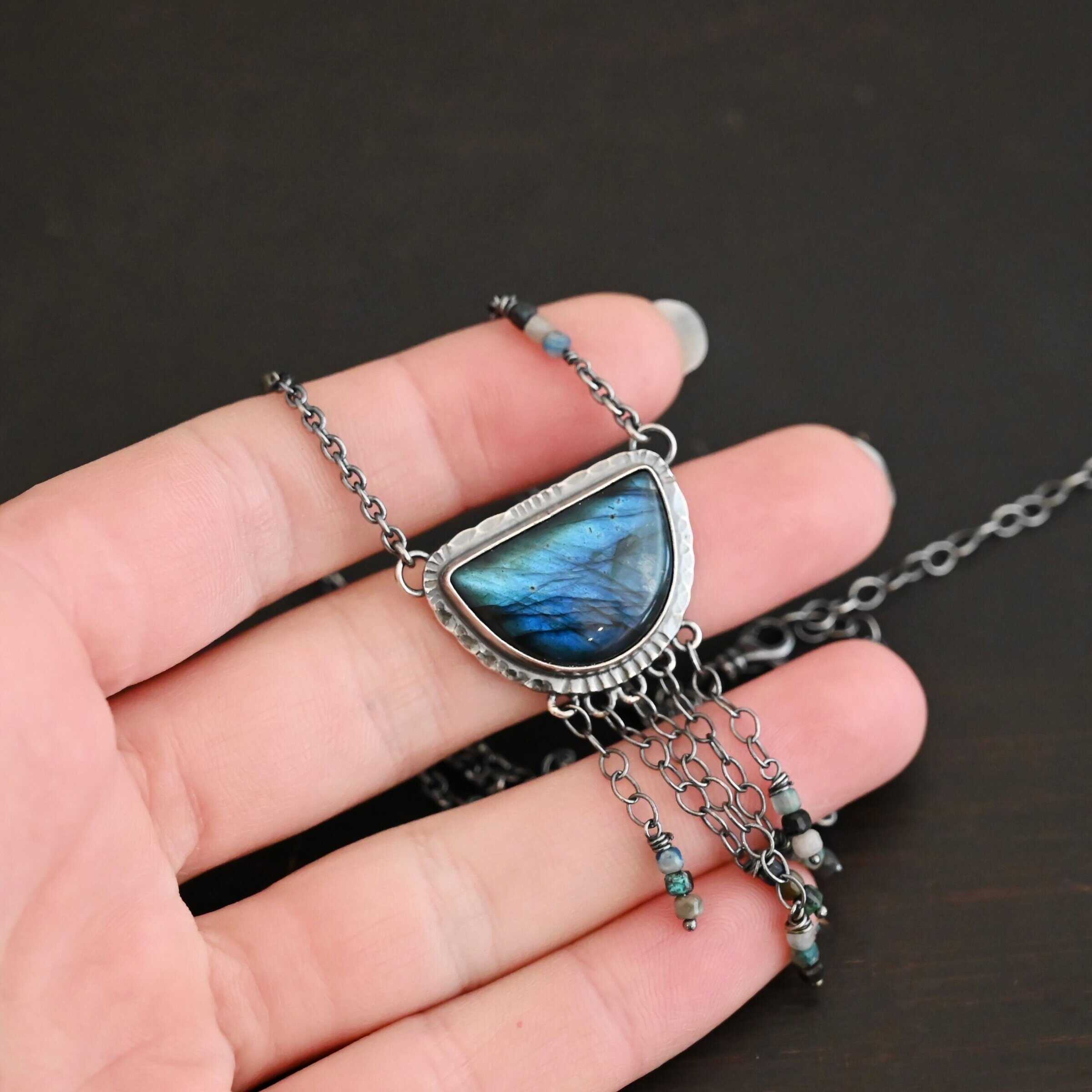 Dream Catcher Necklace Close up of the "Dream Catcher" Necklace. Made out of sterling silver, labradorite, and chrysocolla beads. The labradorite is a half circle shape, with the flat half at the top from where the circle would be cut in half. There are five short strands of silver chain with chrysocolla beads on each end of the chains. The labradorite is set in hammered sterling silver and on each end of the top of the setting is a loop. Attached to the loops are silver chain with chrysocolla beads on them. Necklace is laying against the fingertips of a hand.