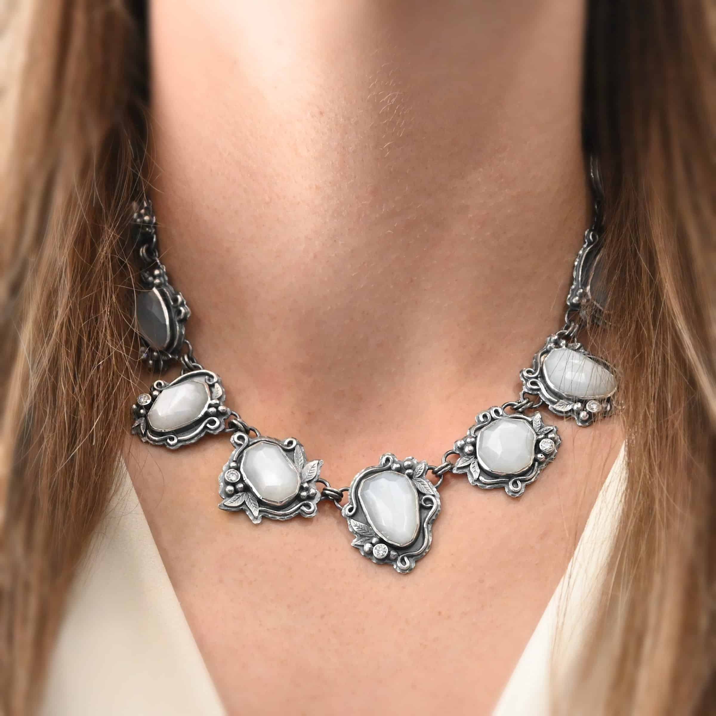 "Among The Stars" Necklace. There are fourteen moonstones that create a collar necklace. Each stone has short, swirling lines of silver around it, as well as silver leaves and silver ball detail. Each moonstone also has a moissanite placed next to it.