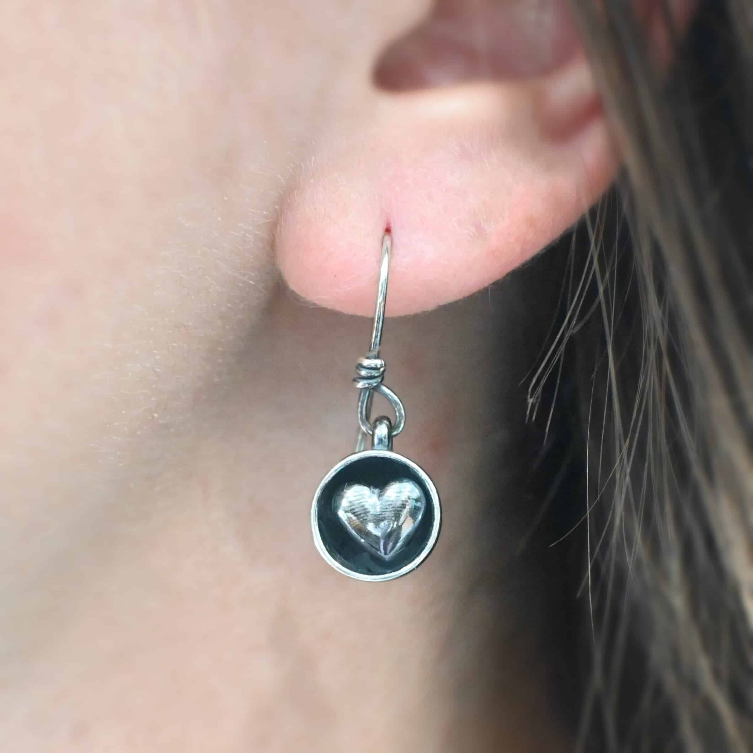 "Cups Of Love" Earrings. Earrings have sterling silver french hook ear wires. Hanging at the bottom of each is a small silver "cup" with a silver heart in the center of it. Earring is shown up close on a model.