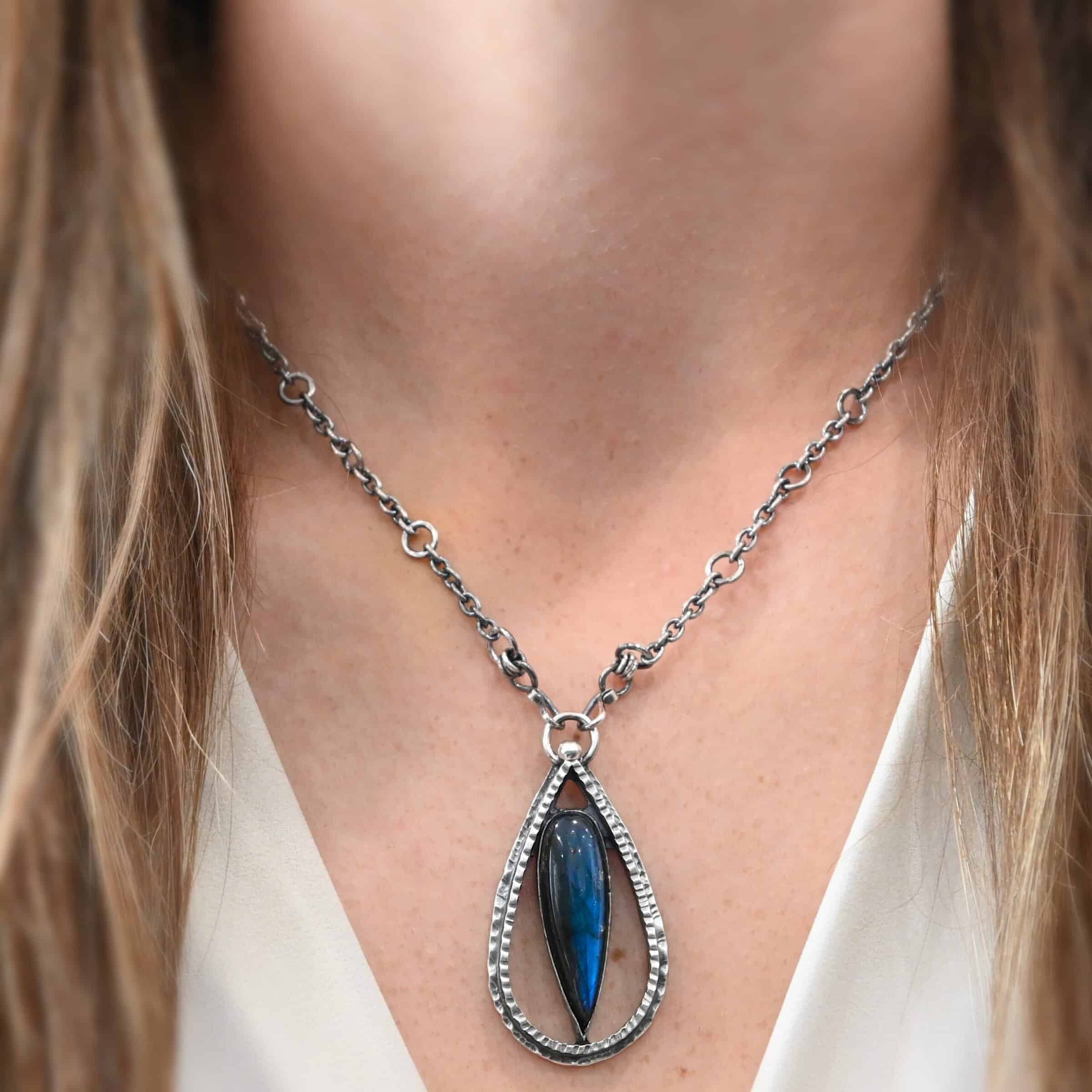 "It Will Rain" Necklace. There is a teardrop of silver pointing upwards. There is a teardrop labradorite pointing downwards in the center of the silver. There is a ball of silver at the top of the silver teardrop. Pendant is on a silver chain.