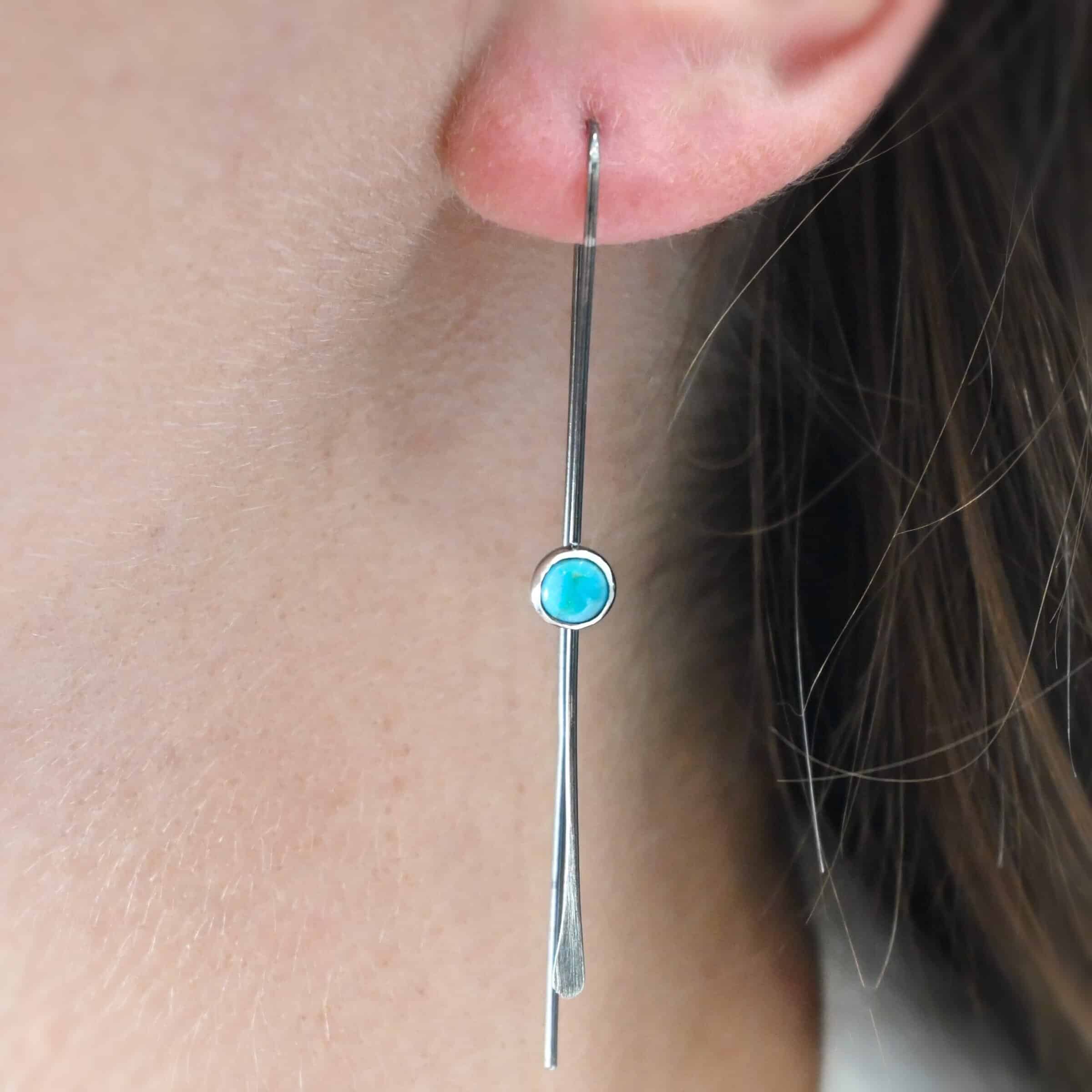 Nimmer Earrings "Nimmer" Earrings. Each earring is a long, thin line of curved silver at the top, with a hammered paddle end. Each earring has a small, circular turquoise in the center of the line on the front. Earring is worn by a model up close.