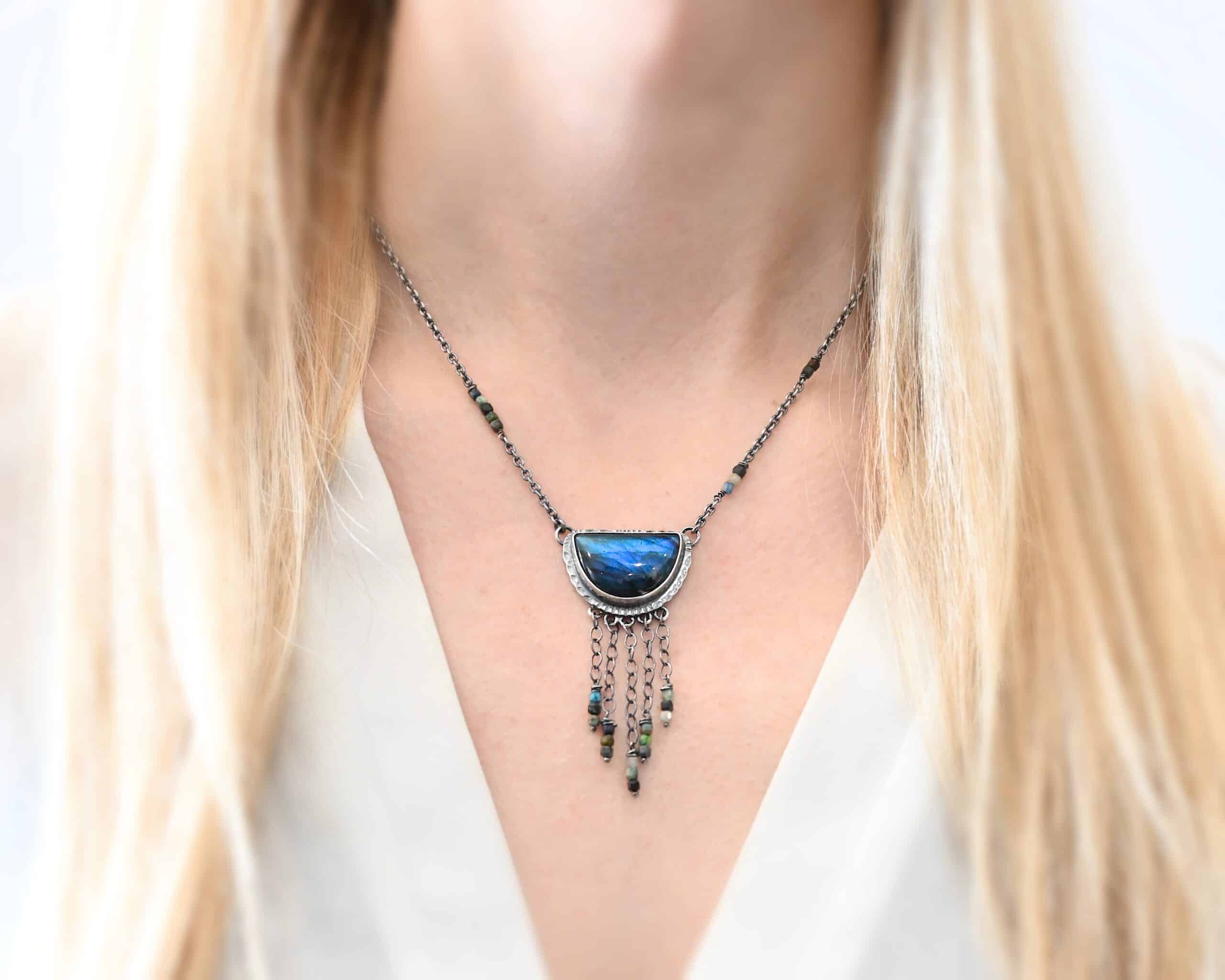 Dream Catcher Necklace Close up of the "Dream Catcher" Necklace. Made out of sterling silver, labradorite, and chrysocolla beads. The labradorite is a half circle shape, with the flat half at the top from where the circle would be cut in half. There are five short strands of silver chain with chrysocolla beads on each end of the chains. The labradorite is set in hammered sterling silver and on each end of the top of the setting is a loop. Attached to the loops are silver chain with chrysocolla beads on them. Necklace is worn up close by a model.