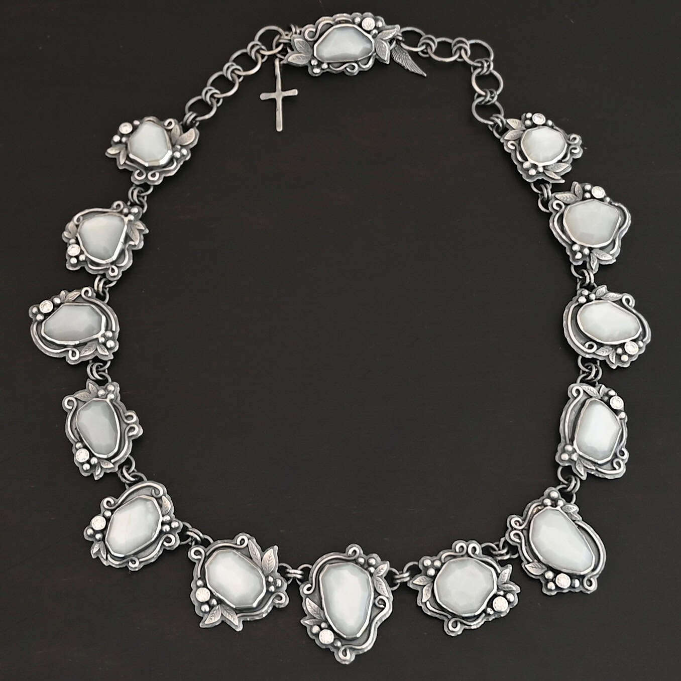 "Among The Stars" Necklace. There are fourteen moonstones that create a collar necklace. Each stone has short, swirling lines of silver around it, as well as silver leaves and silver ball detail. Each moonstone also has a moissanite placed next to it. One of the stones is used as a part of the clasp. There is a cross and angel wing charm on the back of the necklace.