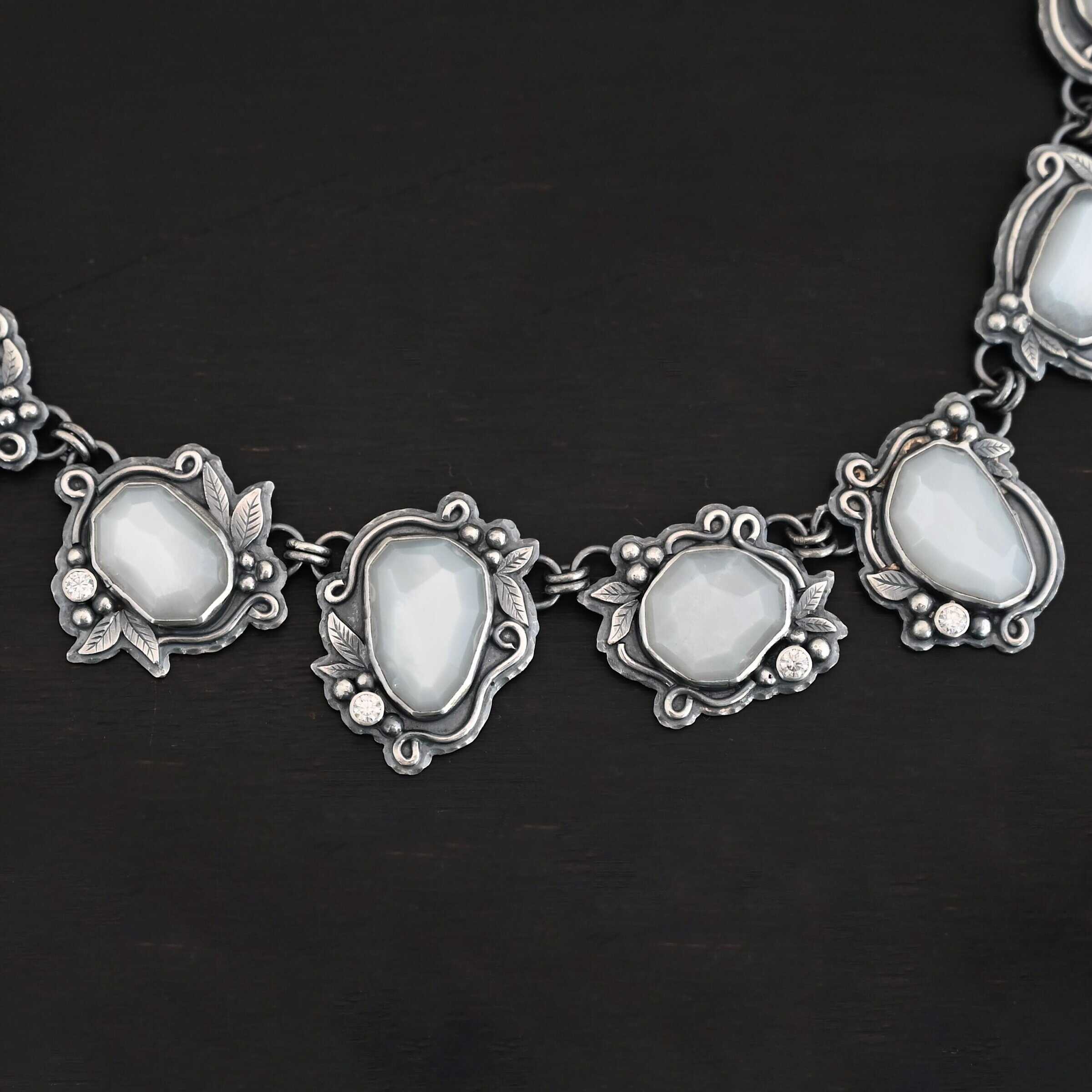 "Among The Stars" Necklace. There are fourteen moonstones that create a collar necklace. Each stone has short, swirling lines of silver around it, as well as silver leaves and silver ball detail. Each moonstone also has a moissanite placed next to it.