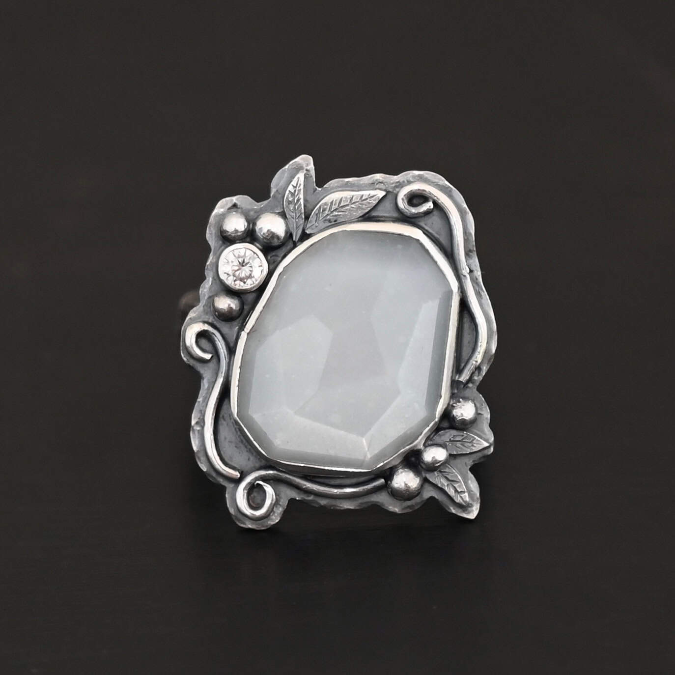 Moonlit Night Ring "Moonlit Night" Ring. There is a rose cut moonstone in the center and a moissanite on the top left side of it. There are silver leaves and short, swirling lines of silver around the stone. There is silver ball detail around the stone as well.