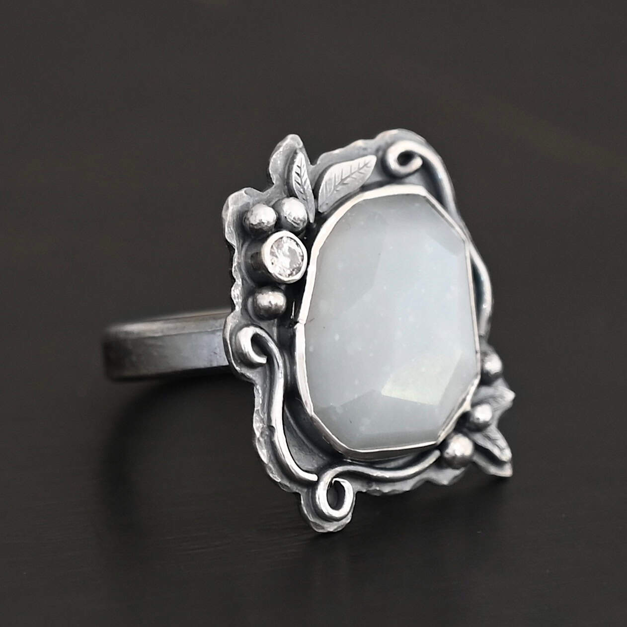 Moonlit Night Ring "Moonlit Night" Ring. There is a rose cut moonstone in the center and a moissanite on the top left side of it. There are silver leaves and short, swirling lines of silver around the stone. There is silver ball detail around the stone as well.