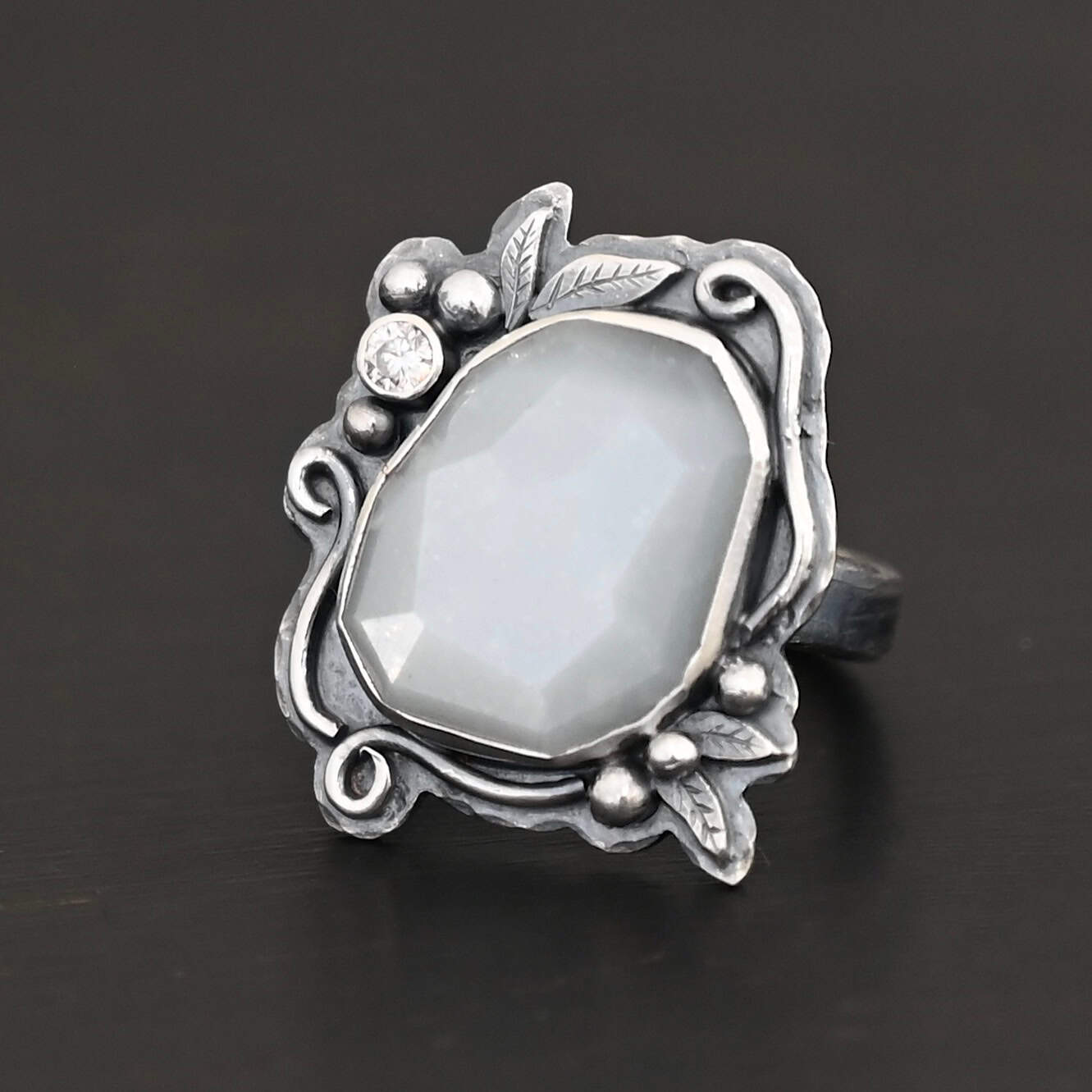 Moonlit Night Ring "Moonlit Night" Ring. There is a rose cut moonstone in the center and a moissanite on the top left side of it. There are silver leaves and short, swirling lines of silver around the stone. There is silver ball detail around the stone as well.