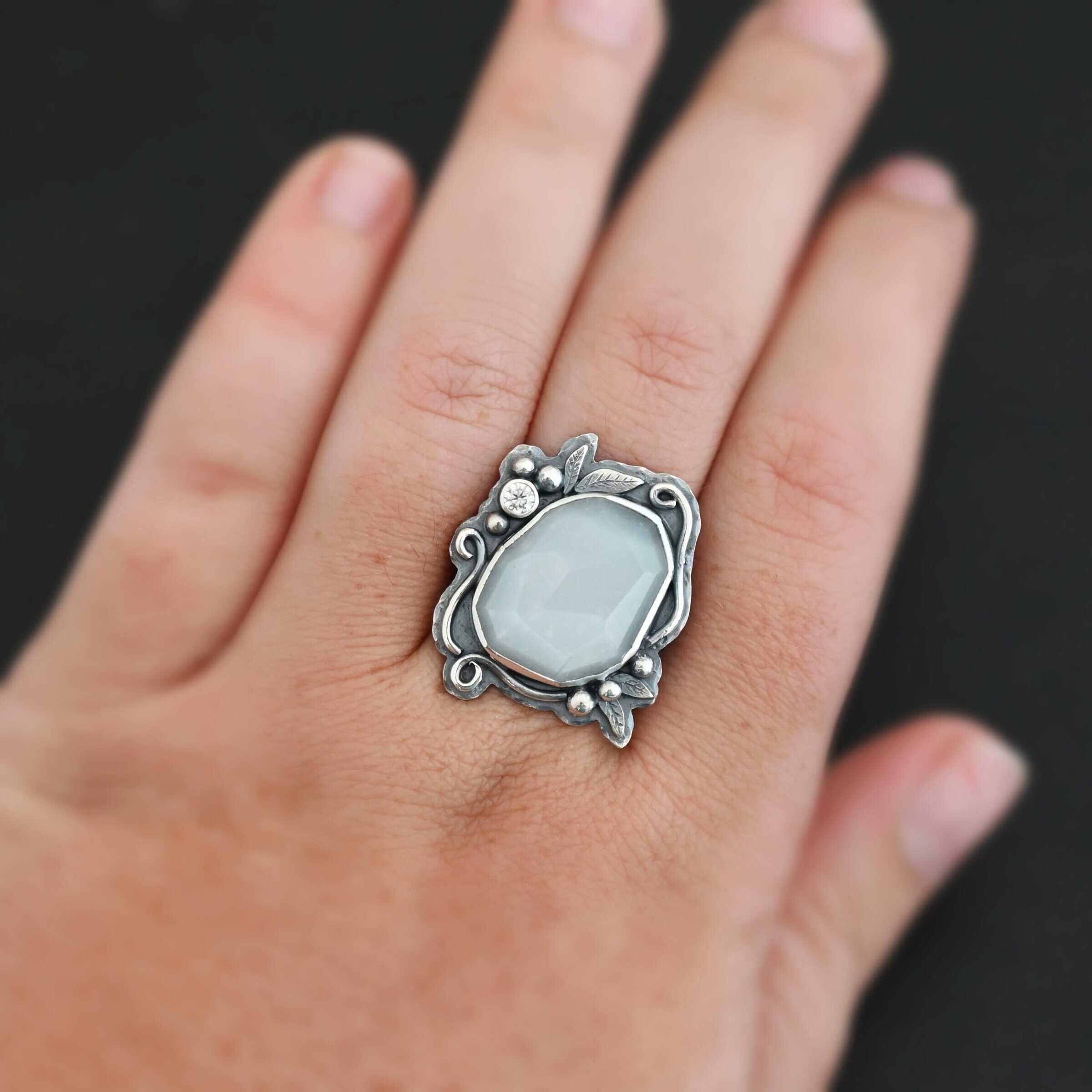 Moonlit Night Ring "Moonlit Night" Ring. There is a rose cut moonstone in the center and a moissanite on the top left side of it. There are silver leaves and short, swirling lines of silver around the stone. There is silver ball detail around the stone as well.