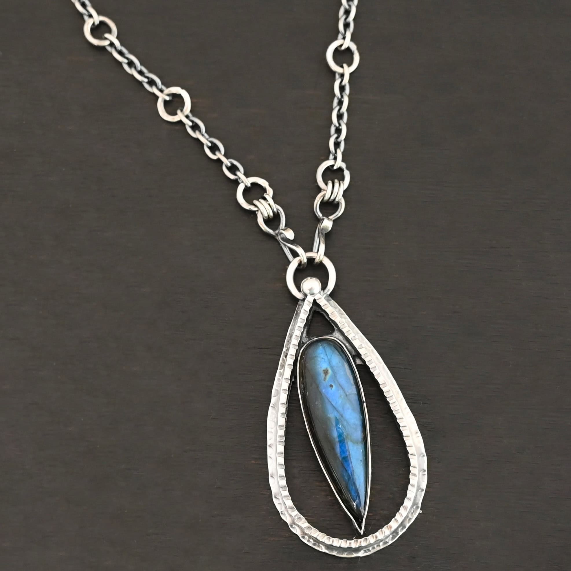 "It Will Rain" Necklace. There is a teardrop of silver pointing upwards. There is a teardrop labradorite pointing downwards in the center of the silver. There is a ball of silver at the top of the silver teardrop. Pendant is on a silver chain.