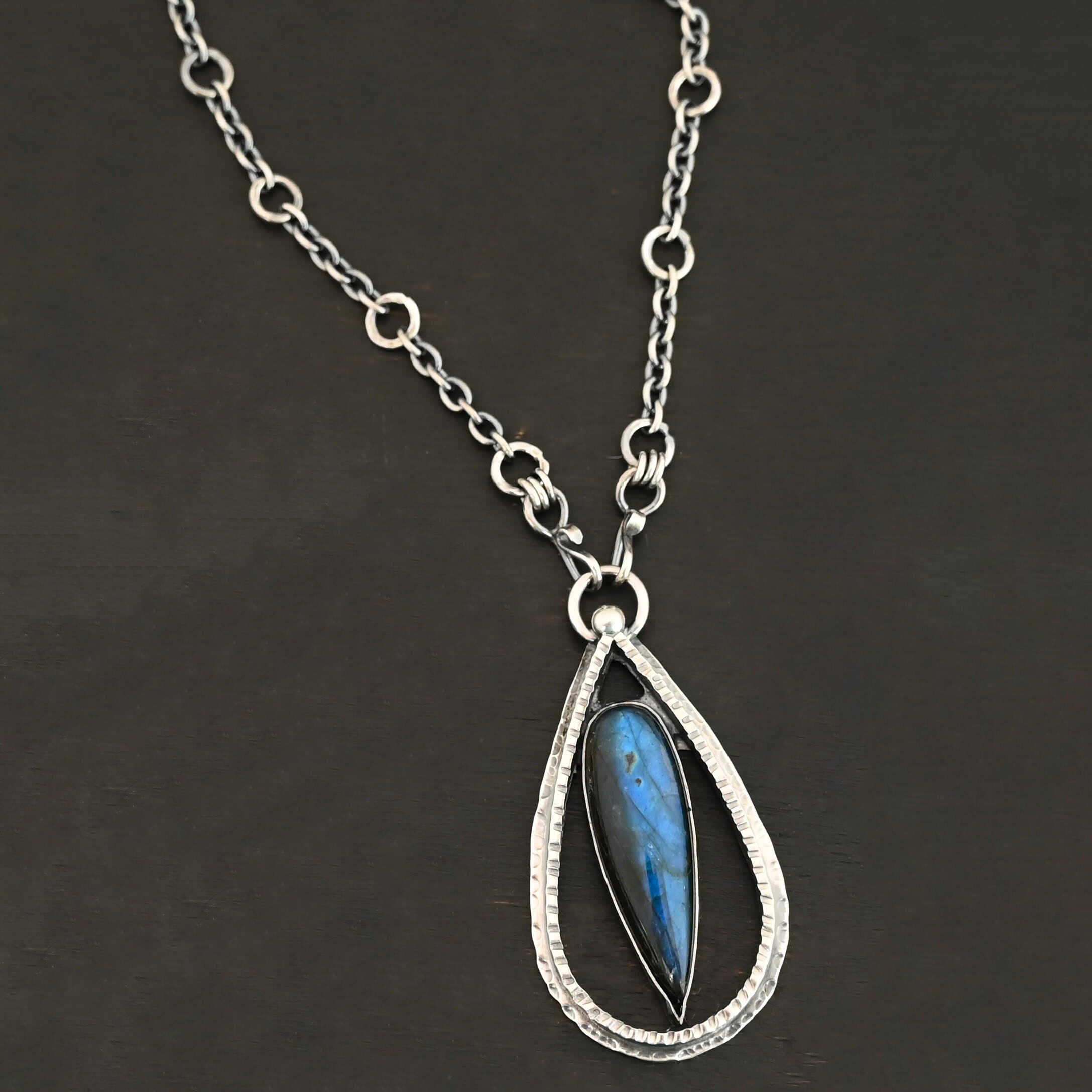 "It Will Rain" Necklace. There is a teardrop of silver pointing upwards. There is a teardrop labradorite pointing downwards in the center of the silver. There is a ball of silver at the top of the silver teardrop. Pendant is on a silver chain.