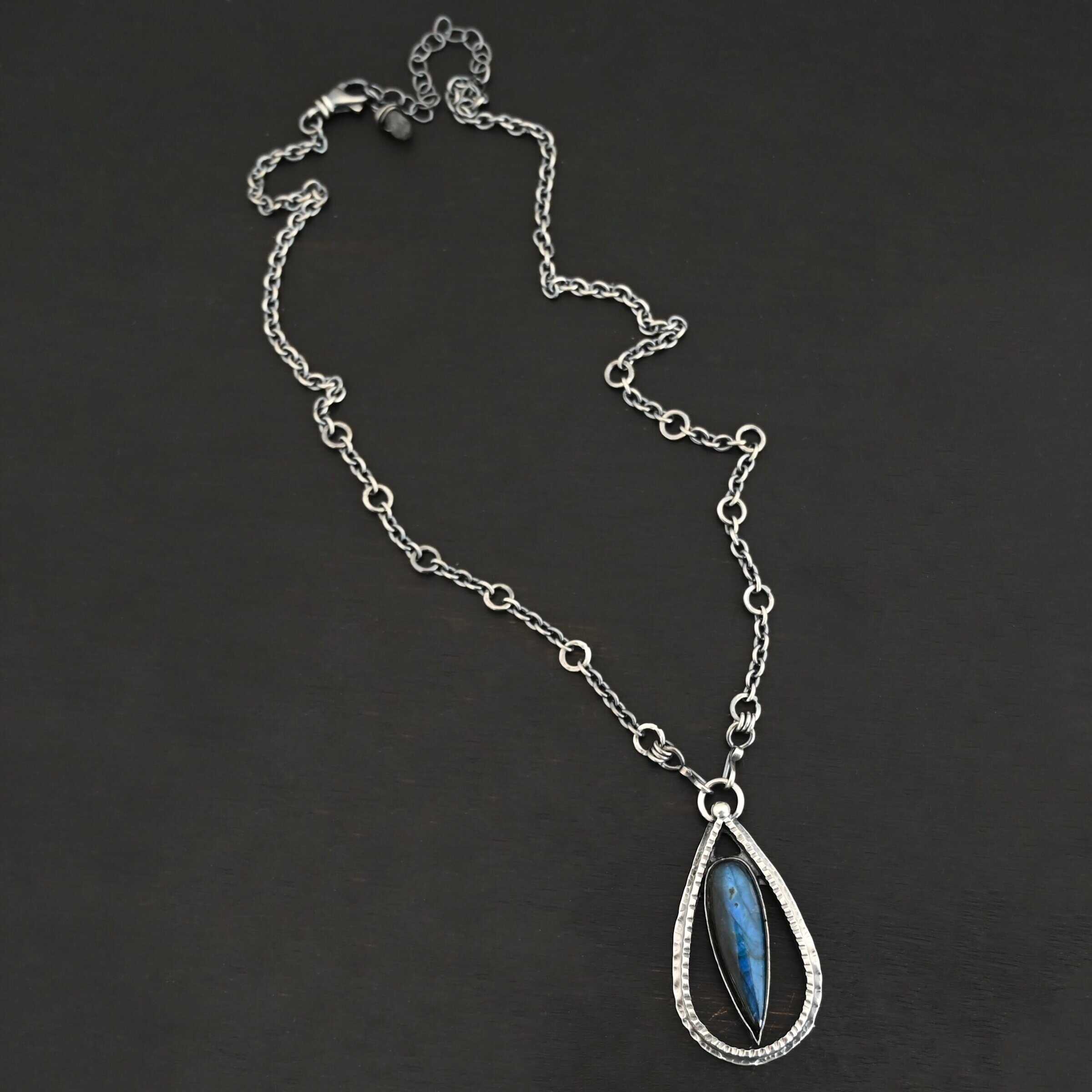 "It Will Rain" Necklace. There is a teardrop of silver pointing upwards. There is a teardrop labradorite pointing downwards in the center of the silver. There is a ball of silver at the top of the silver teardrop. Pendant is on a silver chain.