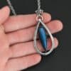 "It Will Rain" Necklace. There is a teardrop of silver pointing upwards. There is a teardrop labradorite pointing downwards in the center of the silver. There is a ball of silver at the top of the silver teardrop. Pendant is on a silver chain.