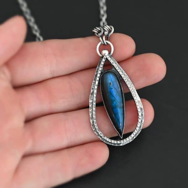"It Will Rain" Necklace. There is a teardrop of silver pointing upwards. There is a teardrop labradorite pointing downwards in the center of the silver. There is a ball of silver at the top of the silver teardrop. Pendant is on a silver chain.