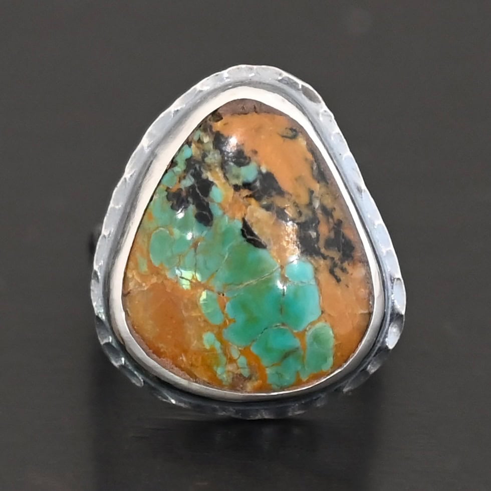 Denver Ring "Denver" Ring. There is a mostly oval turquoise in the center with hammered sterling silver around it. Ring is against a black background.