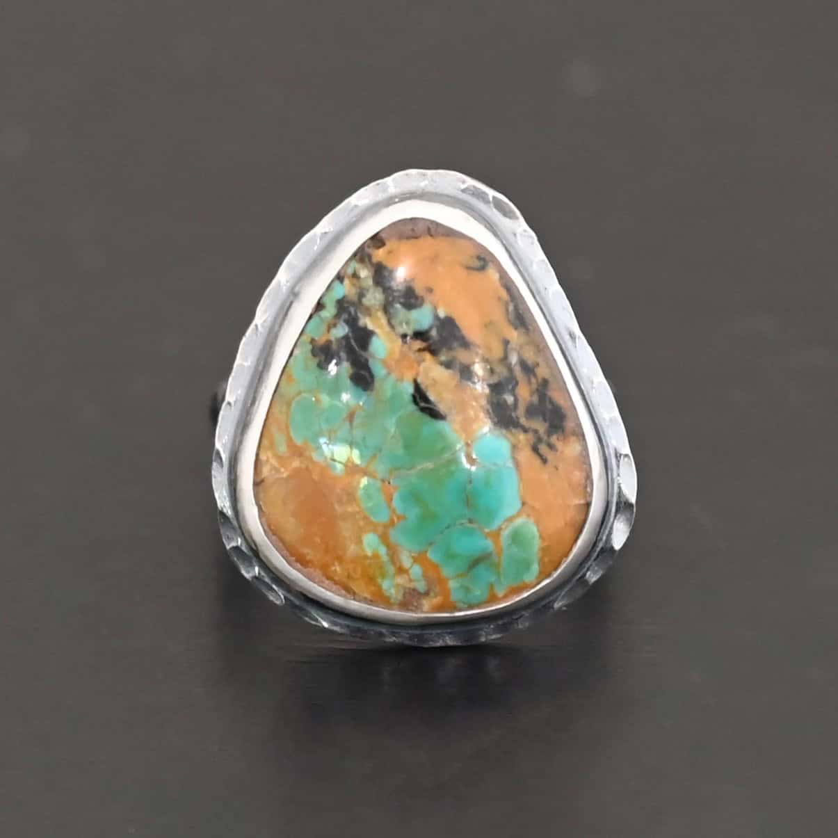 Denver Ring "Denver" Ring. There is a mostly oval turquoise in the center with hammered sterling silver around it. Ring is against a black background.