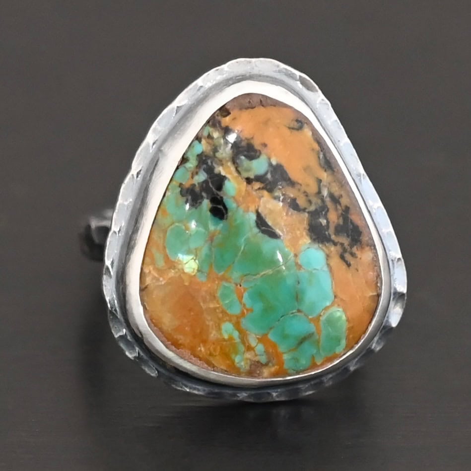 Denver Ring "Denver" Ring. There is a mostly oval turquoise in the center with hammered sterling silver around it. Ring is against a black background.