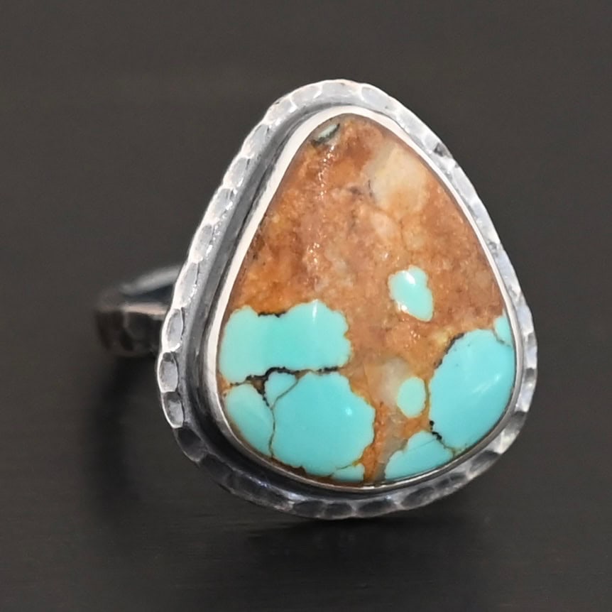 "Arleen" Ring. There is a mostly oval turquoise in the center with hammered sterling silver around it. Ring is set against a black background.