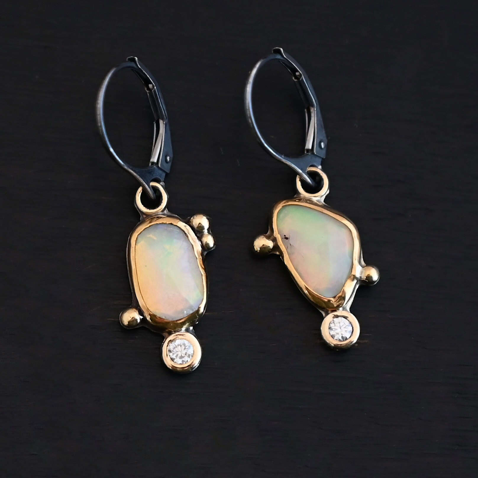 The "Dove Earrings" are made out of sterling silver, gold, opal, and diamond. The opal is the center stone, with the diamond attached directly below the opal. There are three gold dots on the left earring and two on the right, and they are scattered around the center opal. The earrings are set against a dark background.