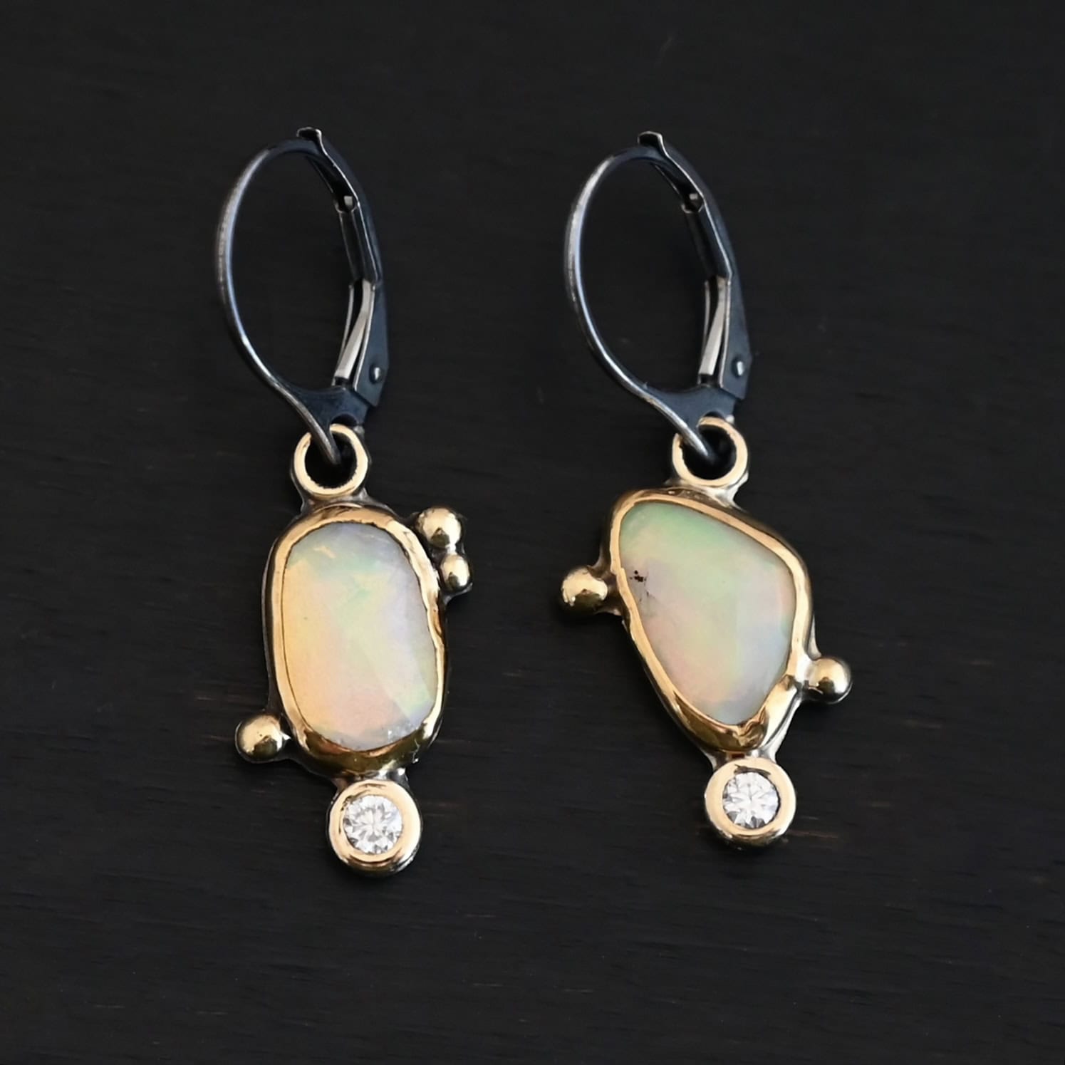 The "Dove Earrings" are made out of sterling silver, gold, opal, and diamond. The opal is the center stone, with the diamond attached directly below the opal. There are three gold dots on the left earring and two on the right, and they are scattered around the center opal. The earrings are set against a dark background.