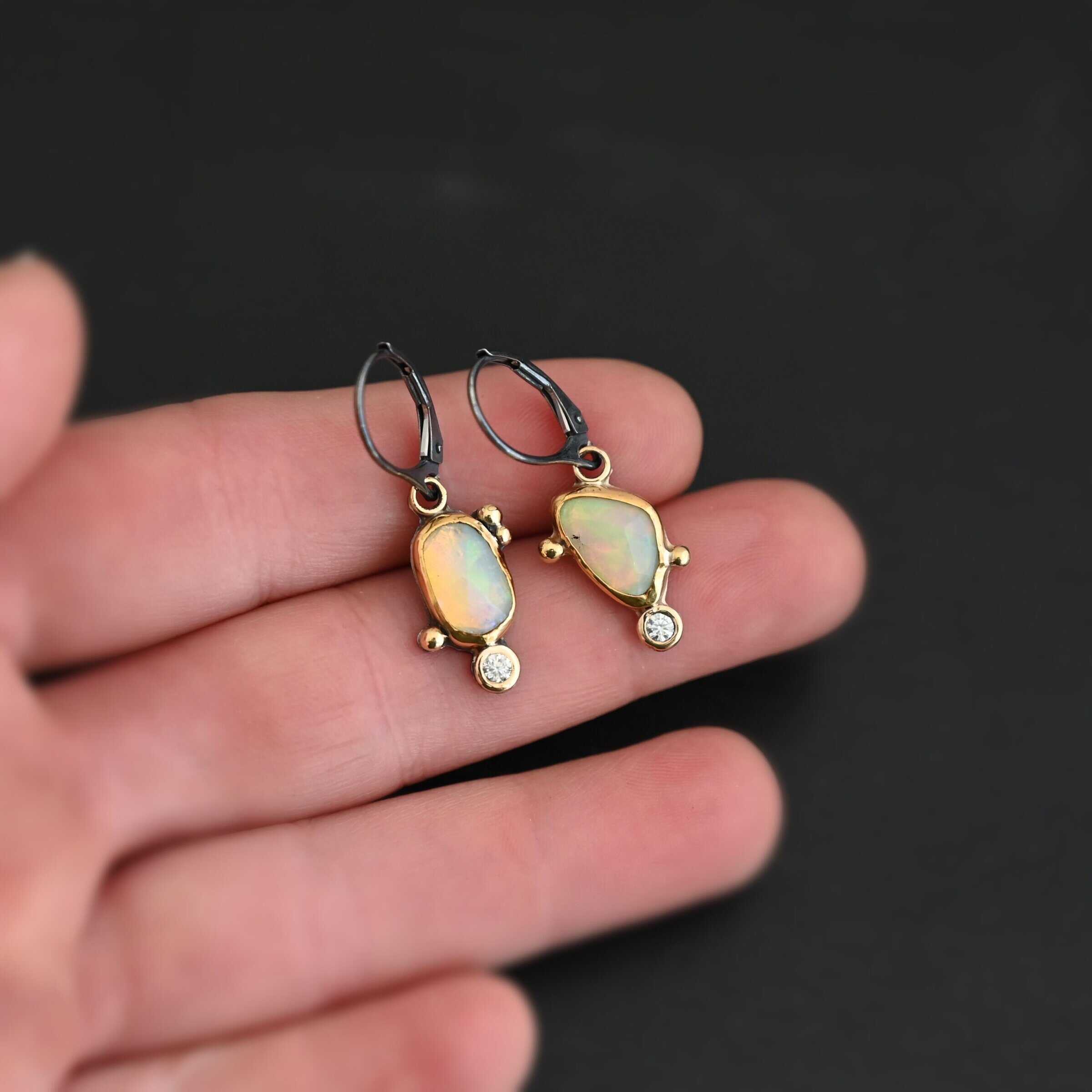 The "Dove Earrings" are made out of sterling silver, gold, opal, and diamond. The opal is the center stone, with the diamond attached directly below the opal. There are three gold dots on the left earring and two on the right, and they are scattered around the center opal.