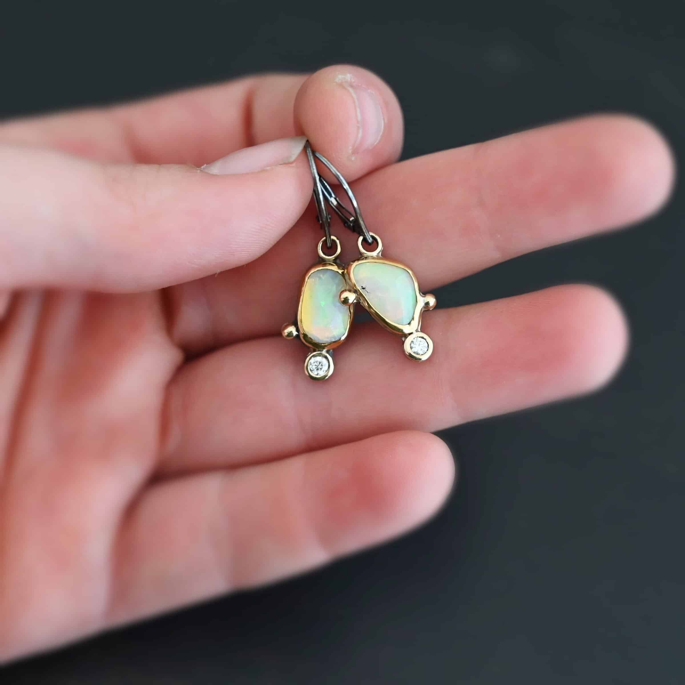The "Dove Earrings" are made out of sterling silver, gold, opal, and diamond. The opal is the center stone, with the diamond attached directly below the opal. There are three gold dots on the left earring and two on the right, and they are scattered around the center opal.