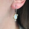 The "Dove Earrings" are made out of sterling silver, gold, opal, and diamond. The opal is the center stone, with the diamond attached directly below the opal. There are three gold dots on the left earring and two on the right, and they are scattered around the center opal.