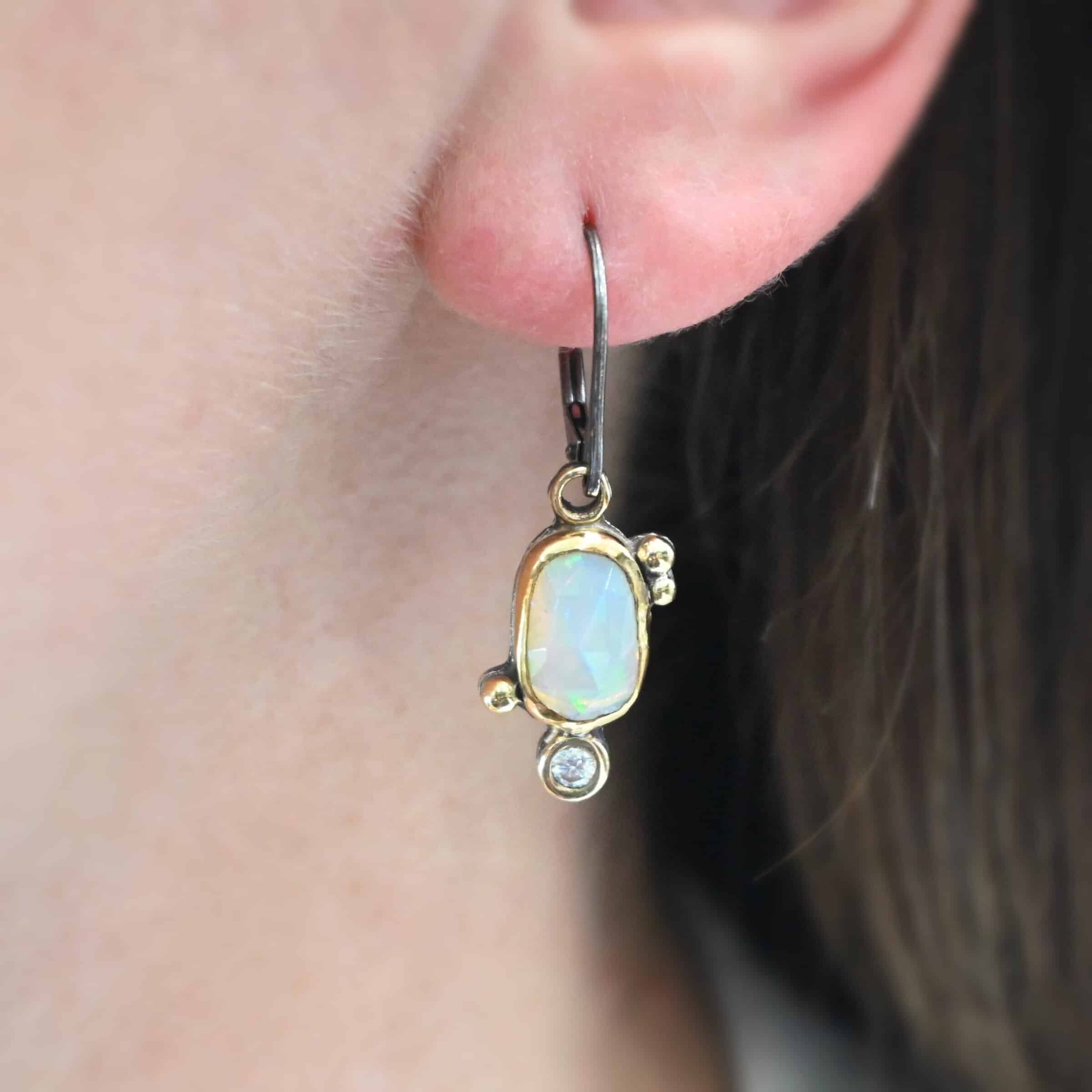 The "Dove Earrings" are made out of sterling silver, gold, opal, and diamond. The opal is the center stone, with the diamond attached directly below the opal. There are three gold dots on the left earring and two on the right, and they are scattered around the center opal.