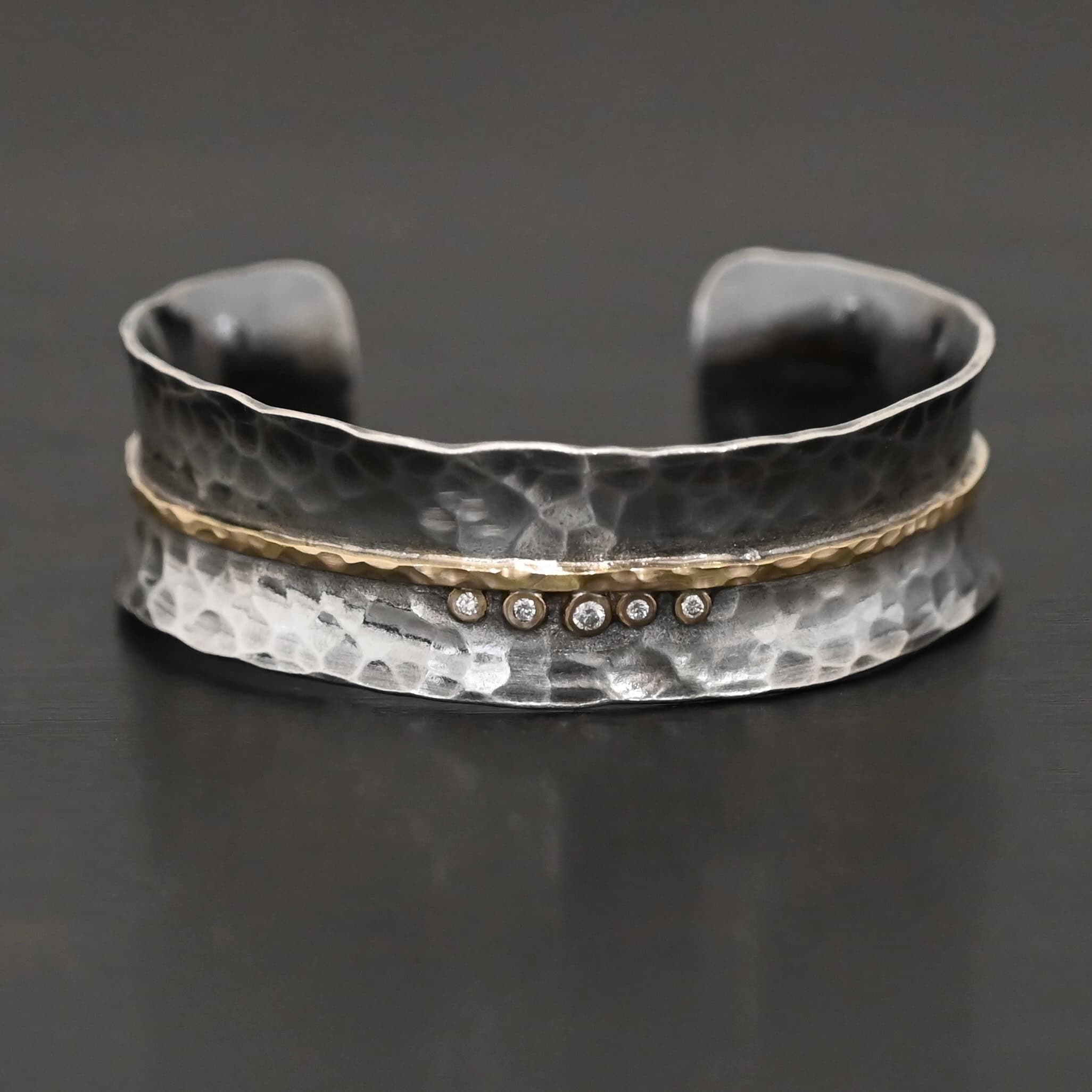Photo of the "Avidity" Cuff. Made out of sterling silver and gold. The cuff is hammered and oxidized. There is a hammered line of gold going straight across the entire cuff through the center. There are five small diamonds set in gold in the center of the cuff, sitting beneath of the line. The dots go in a straight line right next to each other. Cuff is sitting against a dark background.