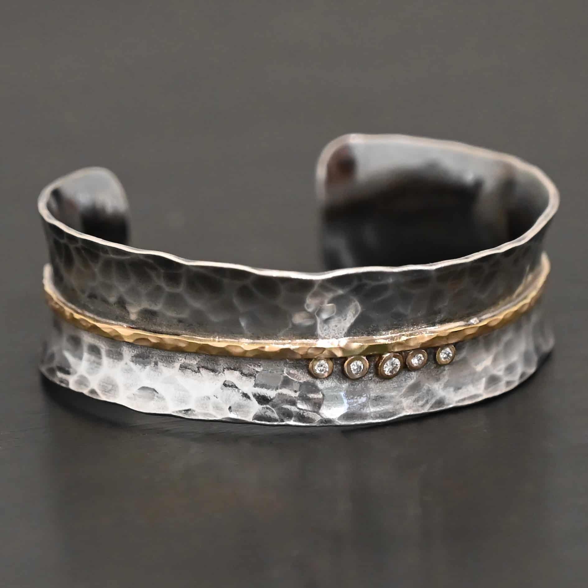 Photo of the "Avidity" Cuff. Made out of sterling silver and gold. The cuff is hammered and oxidized. There is a hammered line of gold going straight across the entire cuff through the center. There are five small diamonds set in gold in the center of the cuff, sitting beneath of the line. The dots go in a straight line right next to each other. Cuff is sitting against a dark background.