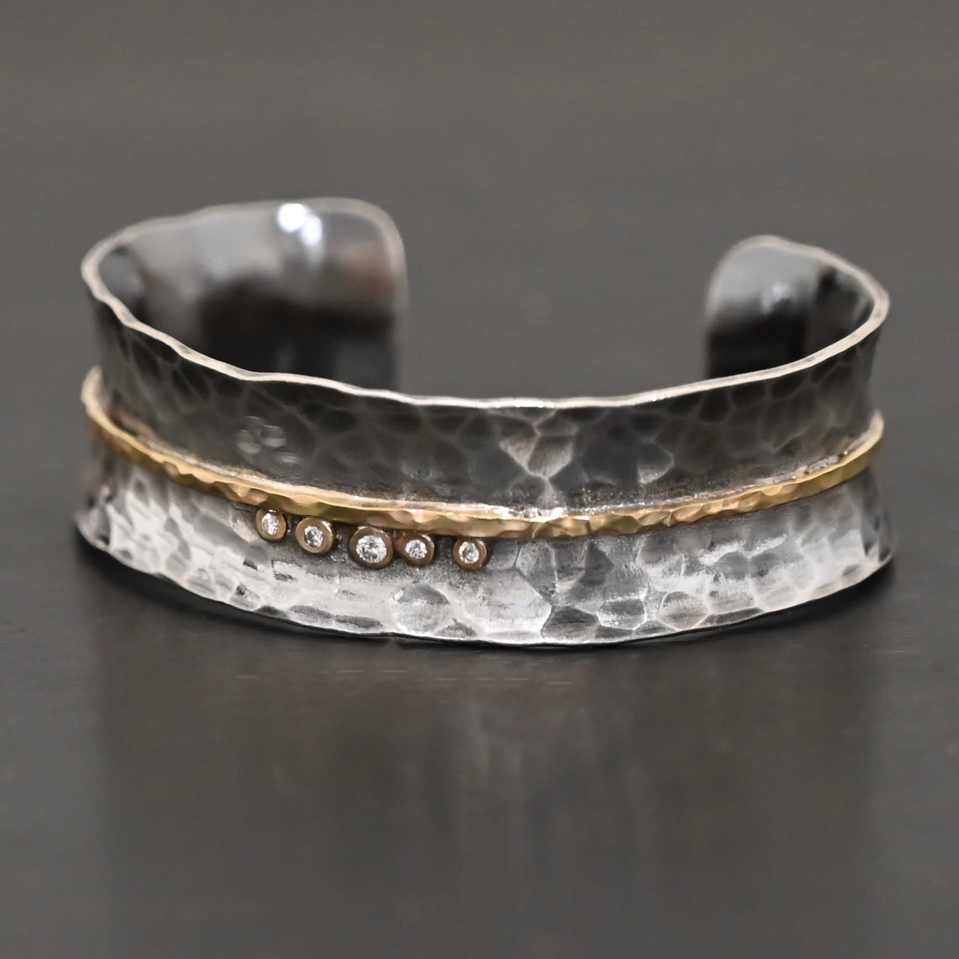 Photo of the "Avidity" Cuff. Made out of sterling silver and gold. The cuff is hammered and oxidized. There is a hammered line of gold going straight across the entire cuff through the center. There are five small diamonds set in gold in the center of the cuff, sitting beneath of the line. The dots go in a straight line right next to each other. Cuff is sitting against a dark background.